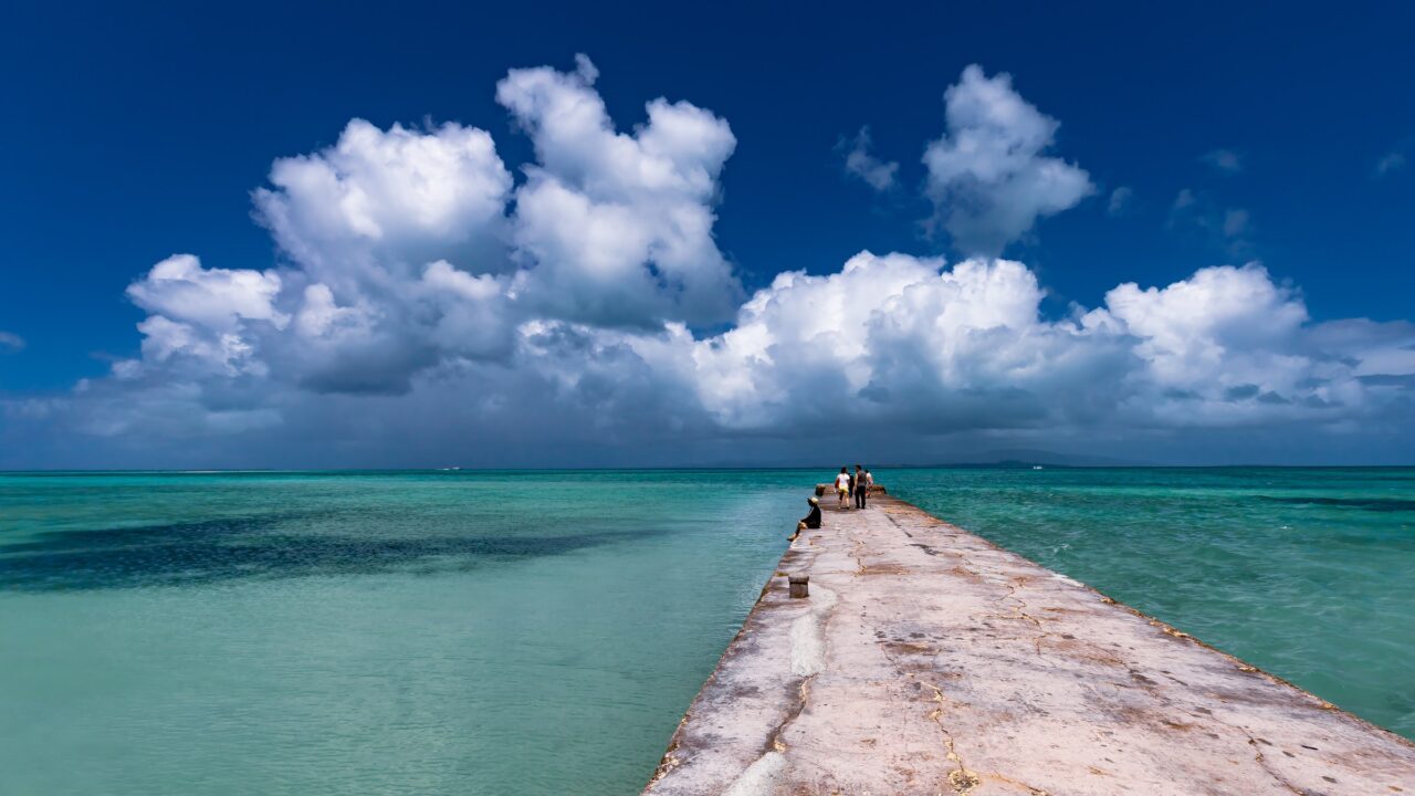A concrete pier running out into a turquoise sea under towering summer clouds