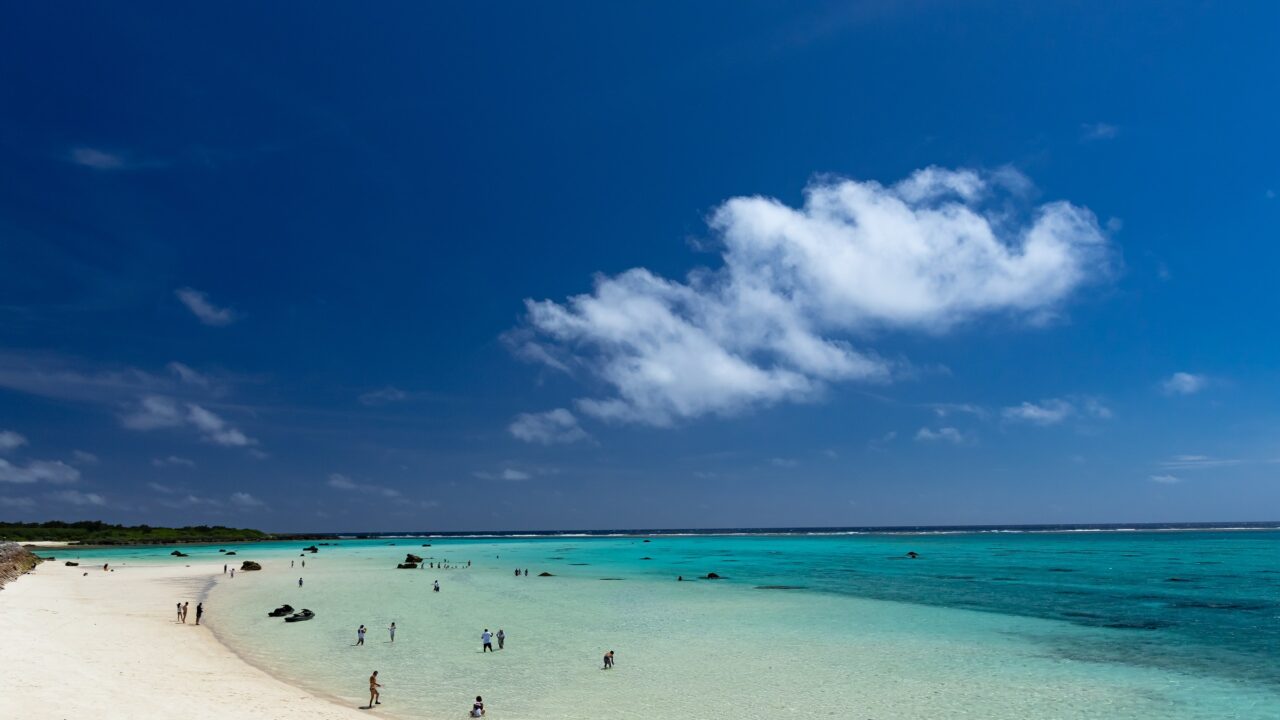 A curve of white sand beach with a few people swimming in clear shallow water
